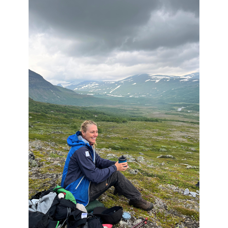 Woman sitting on a mountain slope in hiking gear, holding a cup, with a vast green valley and snow-dotted mountains in the background under a cloudy sky.