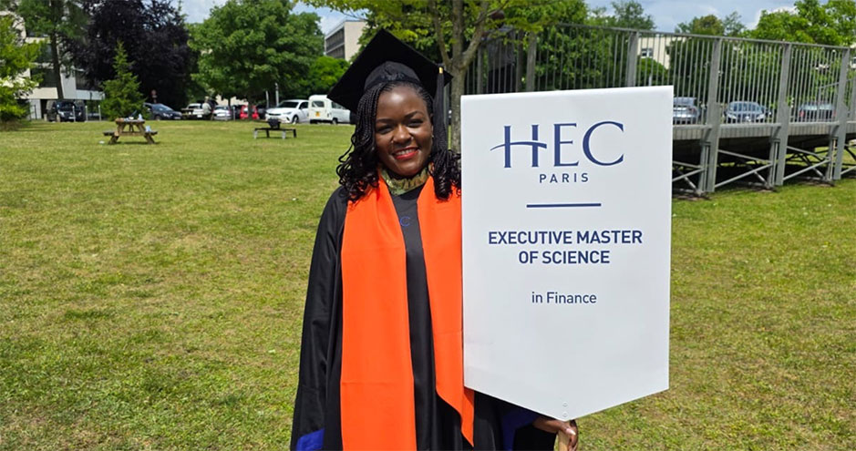 Helen Arrey wearing a black and orange graduation gown, smiling while holding her EMiF diploma in front of an HEC Paris Executive MSc in Finance banner on the campus lawn