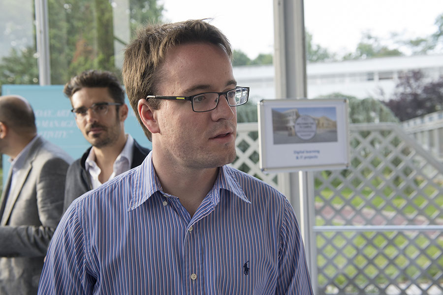 A man in a striped blue shirt wearing glasses is engaged in conversation at an outdoor professional event, with other attendees visible in the background.
