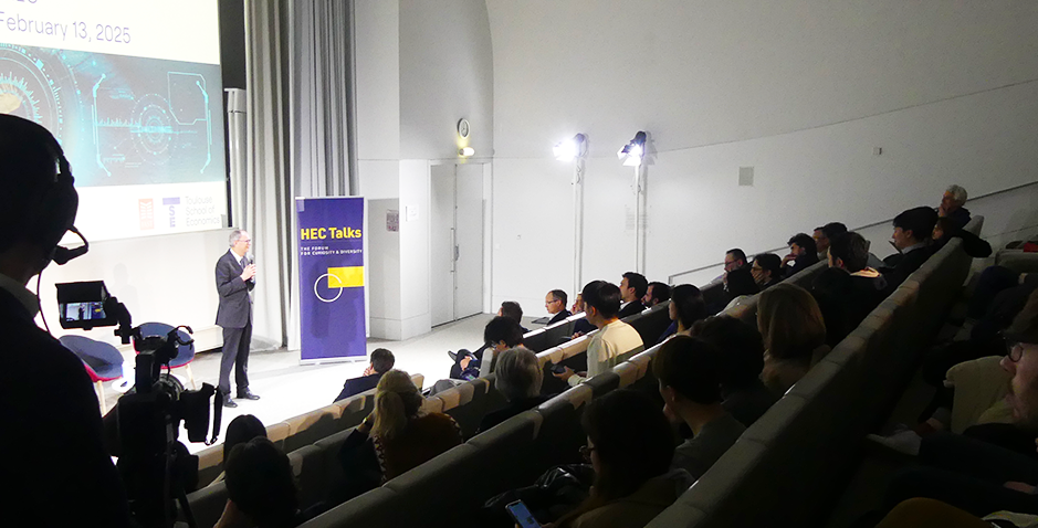 jean tirole presenting at an HEC Talks conference at HEC Paris, addressing an attentive audience in an auditorium, with live video recording taking place.