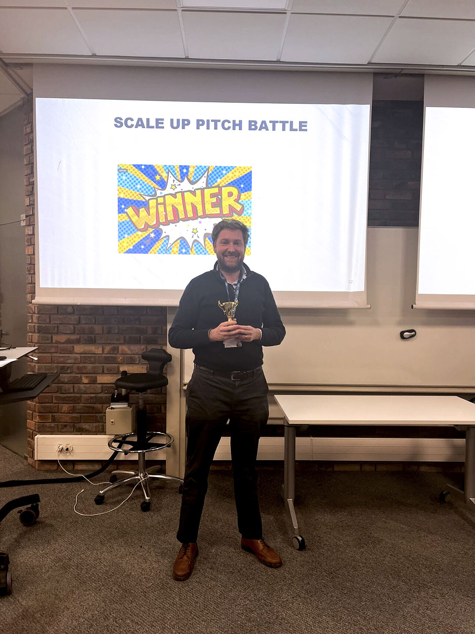 Man standing in front of a large projection screen displaying the title "Scale Up Pitch Battle" and showing a "Winner" graphic