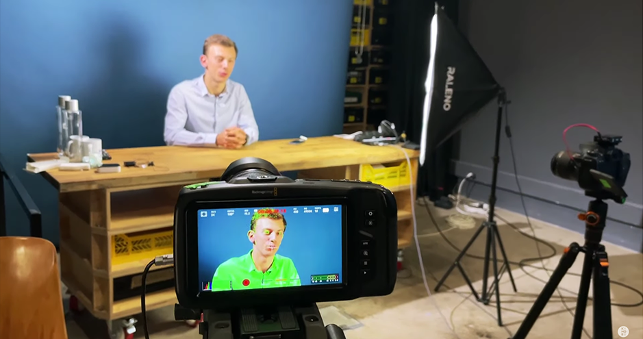 Une personne est assise derrière une table en bois avec un fond bleu. En avant-plan, on voit un écran de caméra montrant la même personne en train de parler. Il y a un éclairage de studio et une autre caméra sur trépied à droite.