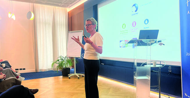 A woman stands in front of a projected slide, gesturing with her hands while addressing a seated audience in a bright conference room