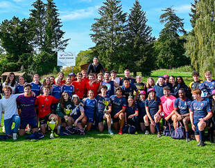 Photo de groupe des équipes gagnantes de la Natixis Rugby Cup posant sur le terrain avec leurs trophées, entourées de nature et d'arbres en arrière-plan