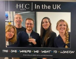 Five smiling people posing with a photo frame that reads HEC Paris In the UK - The One Where We Went to London' at an event in London