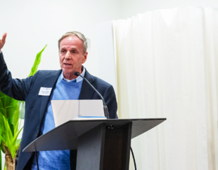 Michael Posner speaking at a podium during a conference, wearing a suit and blue sweater, with plants in the background