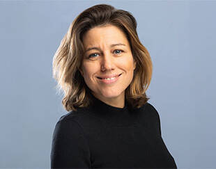 Portrait de Caroline Faillet, souriante, de trois quarts face, portant un pull noir à col montant, sur un fond uni bleu-gris.