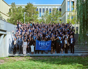Group photo of the September 2025 intake outside holding the HEC Paris MBA flag in front of the S building.