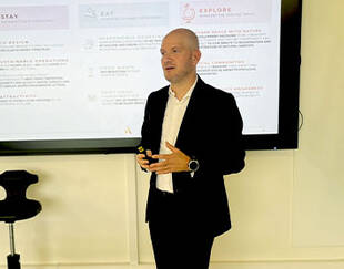 Un homme en costume noir fait une présentation devant un écran qui affiche des points sur l'éco-conception, les opérations durables, et l'expérience client ("STAY, EAT, EXPLORE")