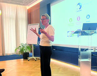 A woman stands in front of a projected slide, gesturing with her hands while addressing a seated audience in a bright conference room