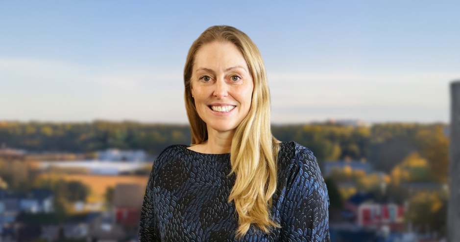 Woman smiling in front of a blurred cityscape, wearing a dark patterned blouse.