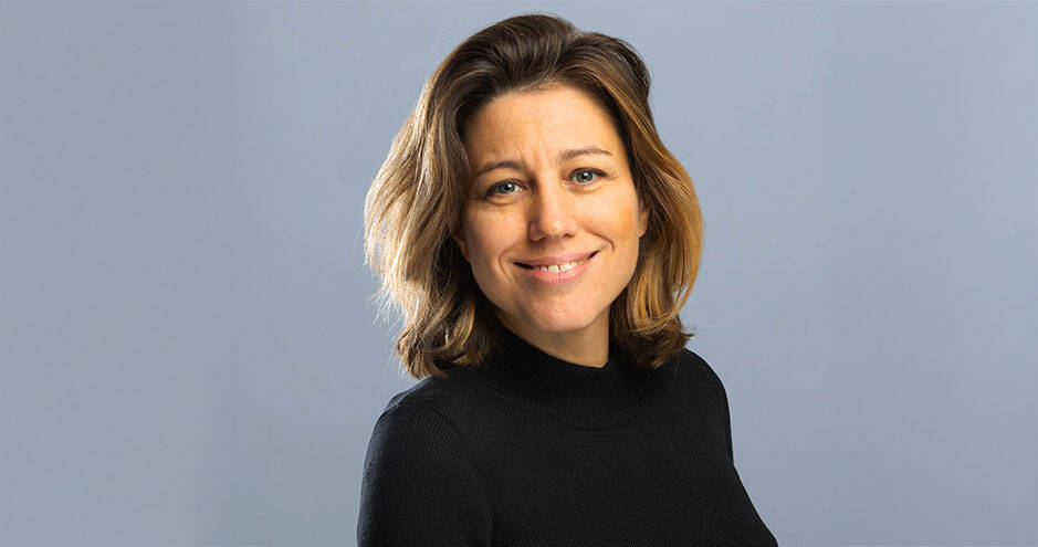 Portrait de Caroline Faillet, souriante, de trois quarts face, portant un pull noir à col montant, sur un fond uni bleu-gris.