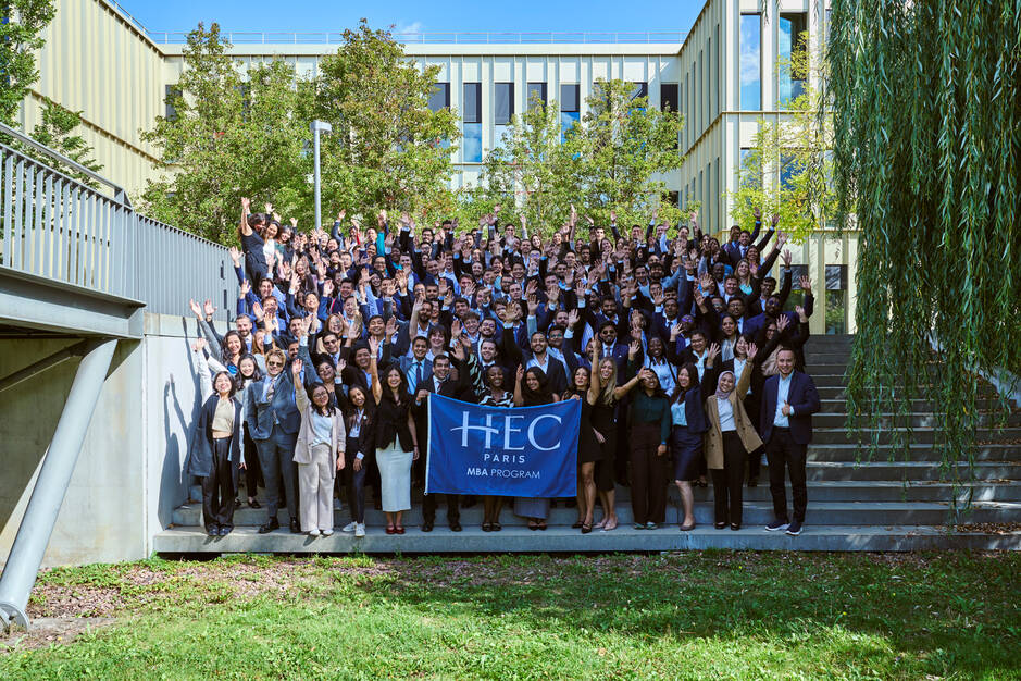 Group photo of the September 2025 intake outside holding the HEC Paris MBA flag in front of the S building.