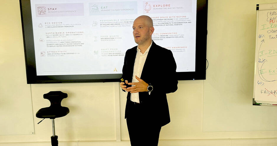 Un homme en costume noir fait une présentation devant un écran qui affiche des points sur l'éco-conception, les opérations durables, et l'expérience client ("STAY, EAT, EXPLORE")
