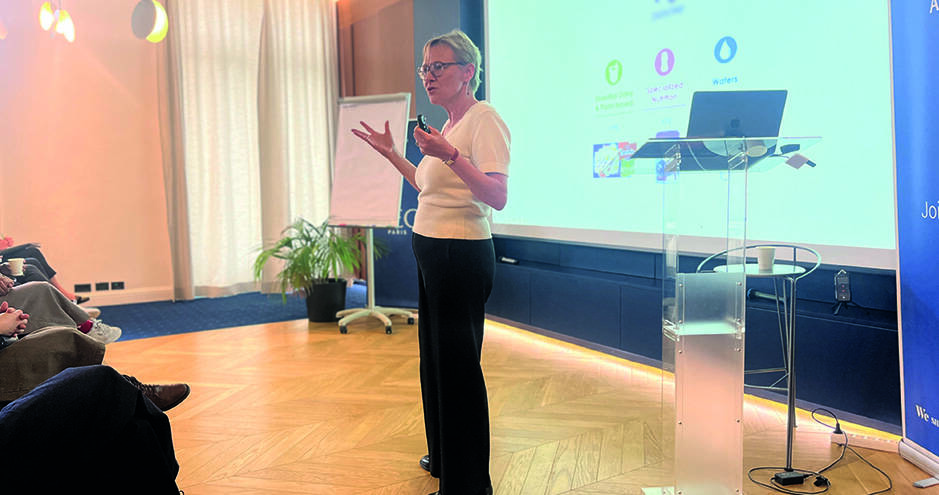 A woman stands in front of a projected slide, gesturing with her hands while addressing a seated audience in a bright conference room